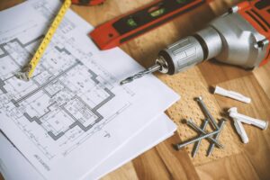 Home renovation planning with blueprint, power drill, screws, anchors, level, and tape measure on wood floor.